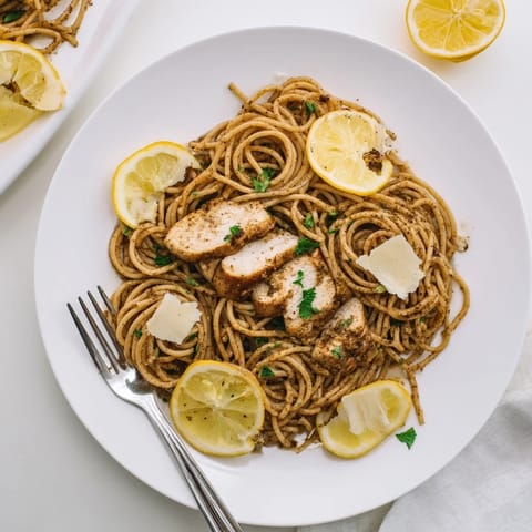 A close-up of brown butter lemon chicken pasta served with lemon wedges and grated Parmesan on a rustic table.